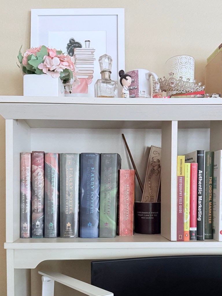 The top shelf has some artwork of a girl holding a lot of books then a small pot of pink hydrangeas. Next to it is a Juicy Couture perfume bottle, a mug, a white candle and a tiara. The second level has the hardbacks of the Harry Potter series, a color changing mug, a wand and the Marauder's map 